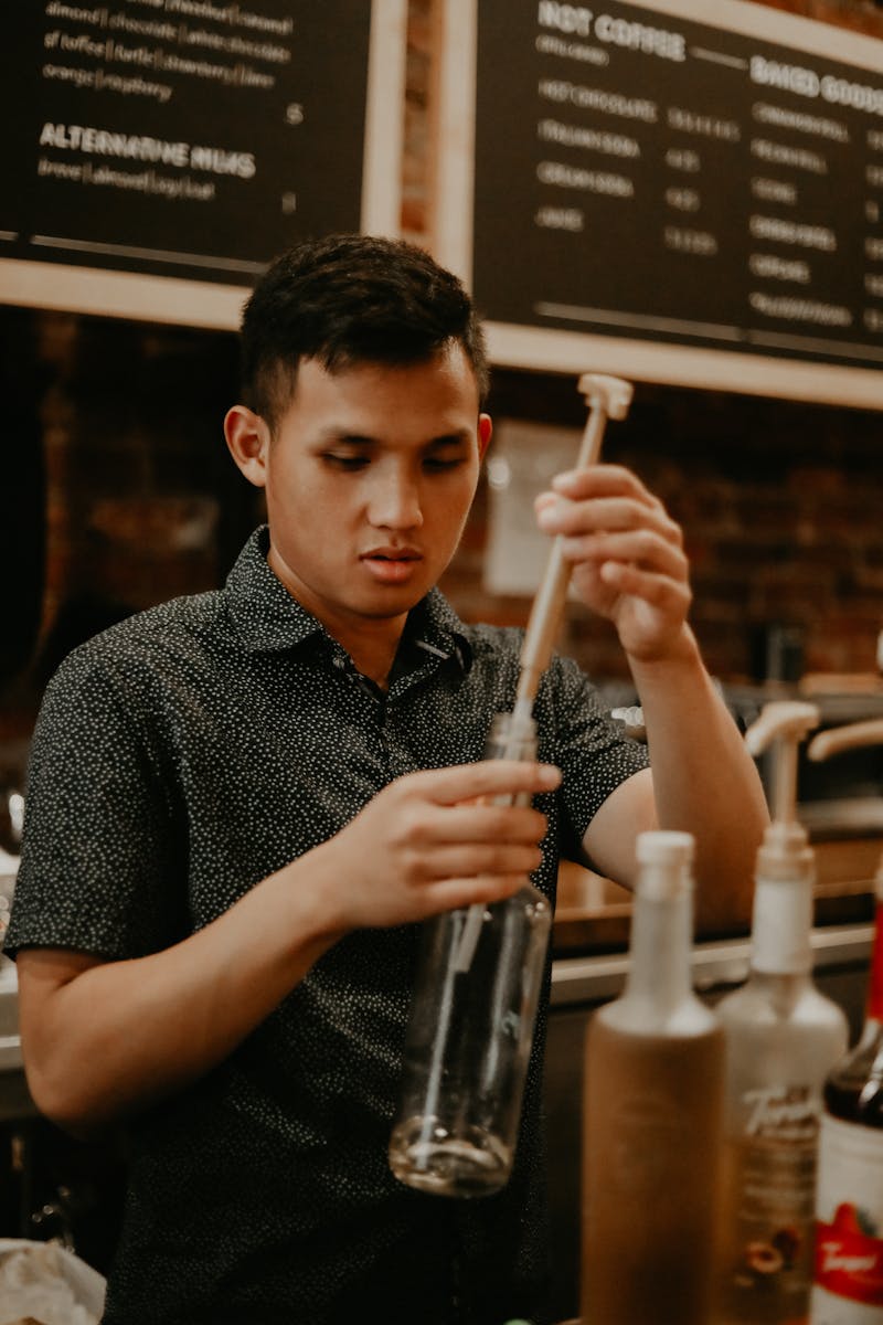 Concentrated ethnic male barista removing pump from syrup bottle while working in modern cafe